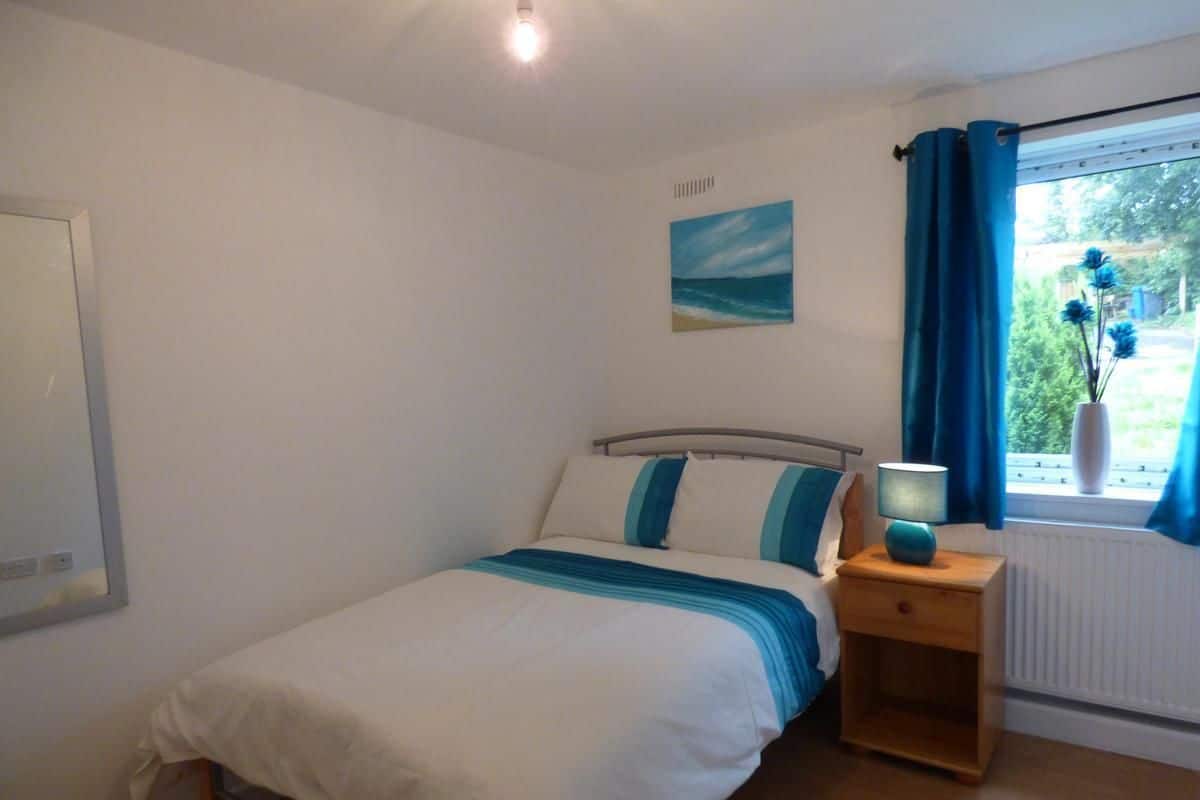 A bedroom with a bed, a nightstand with a lamp, a window with curtains, and a painting on the wall.