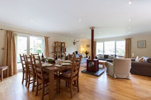 Un vaste espace de vie comprenant une table à manger en bois, des chaises et un canapé. On y trouve également une cheminée, une bibliothèque et une fenêtre avec des rideaux.
