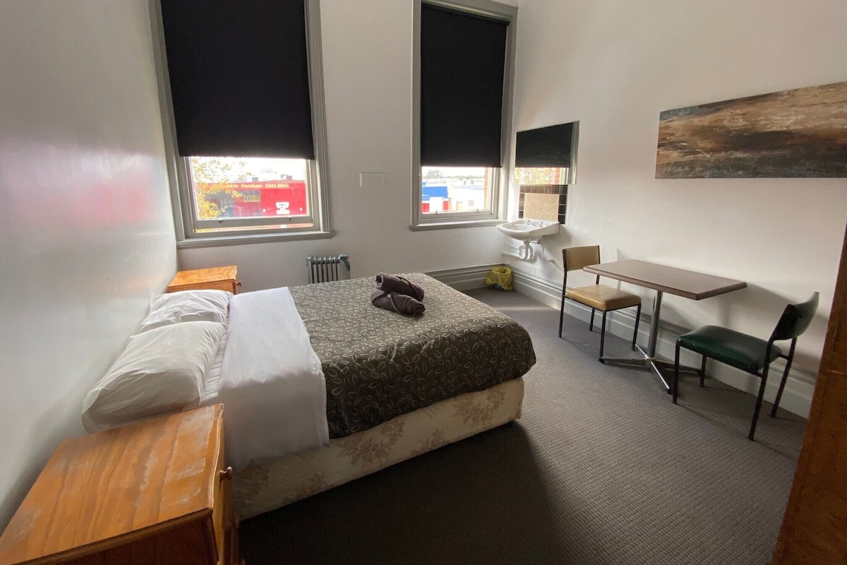 Habitación de hotel con cama, escritorio, silla, radiador y ventana con vista a un edificio.