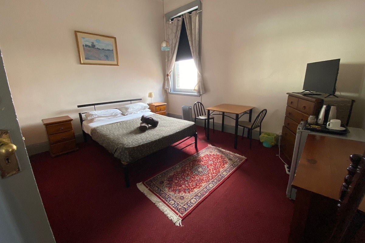 Un dormitorio con cama, escritorio, silla, cómoda, televisor y ventana con cortinas.