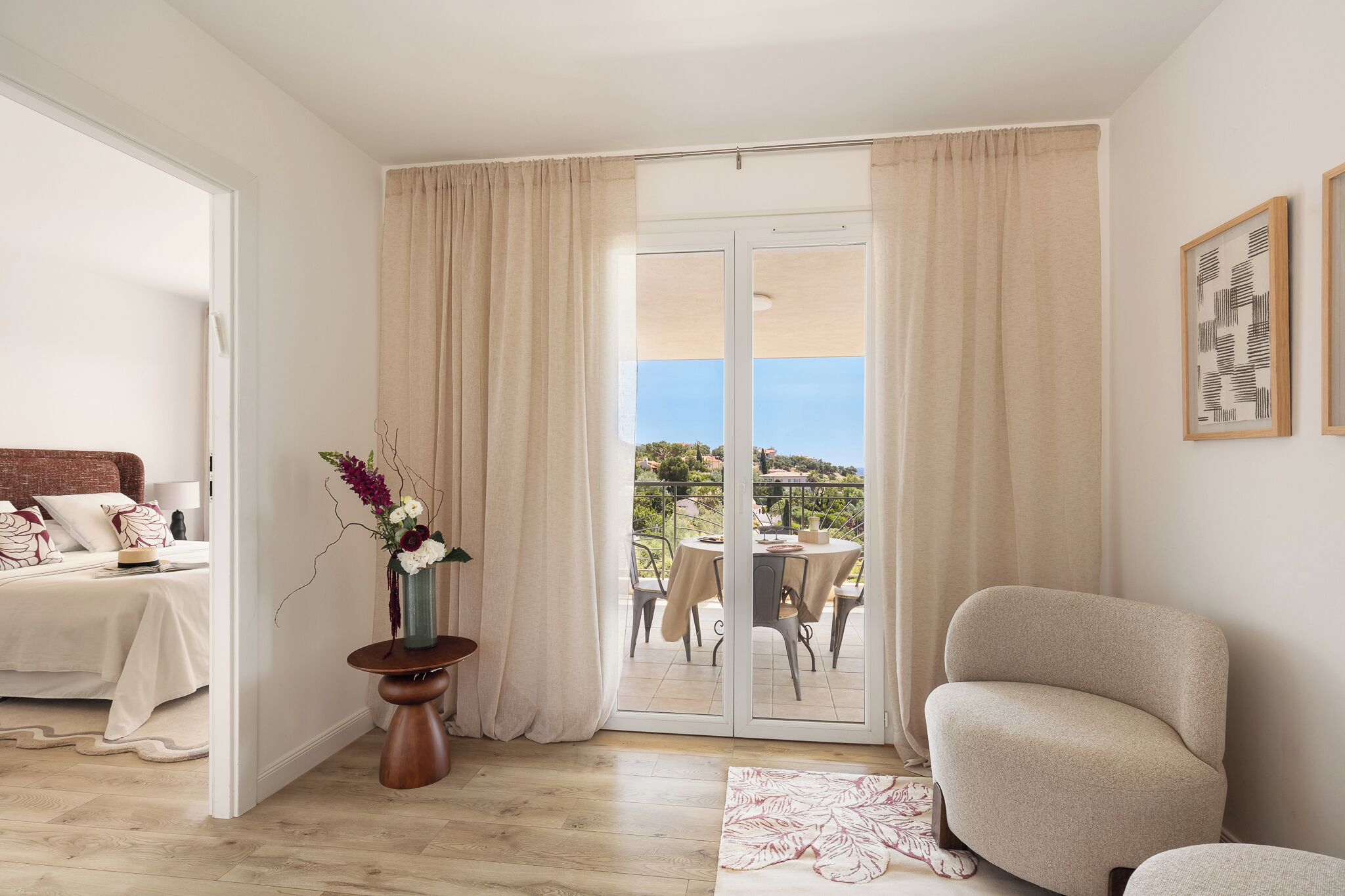 A bedroom with a bed, a bedside table with a vase, and a view of a balcony with a table and chairs.