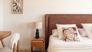 Ett välordnat hotellrum med en säng, kuddar, ett sängbord med en lampa, en stol och ett inramat konstverk på väggen.