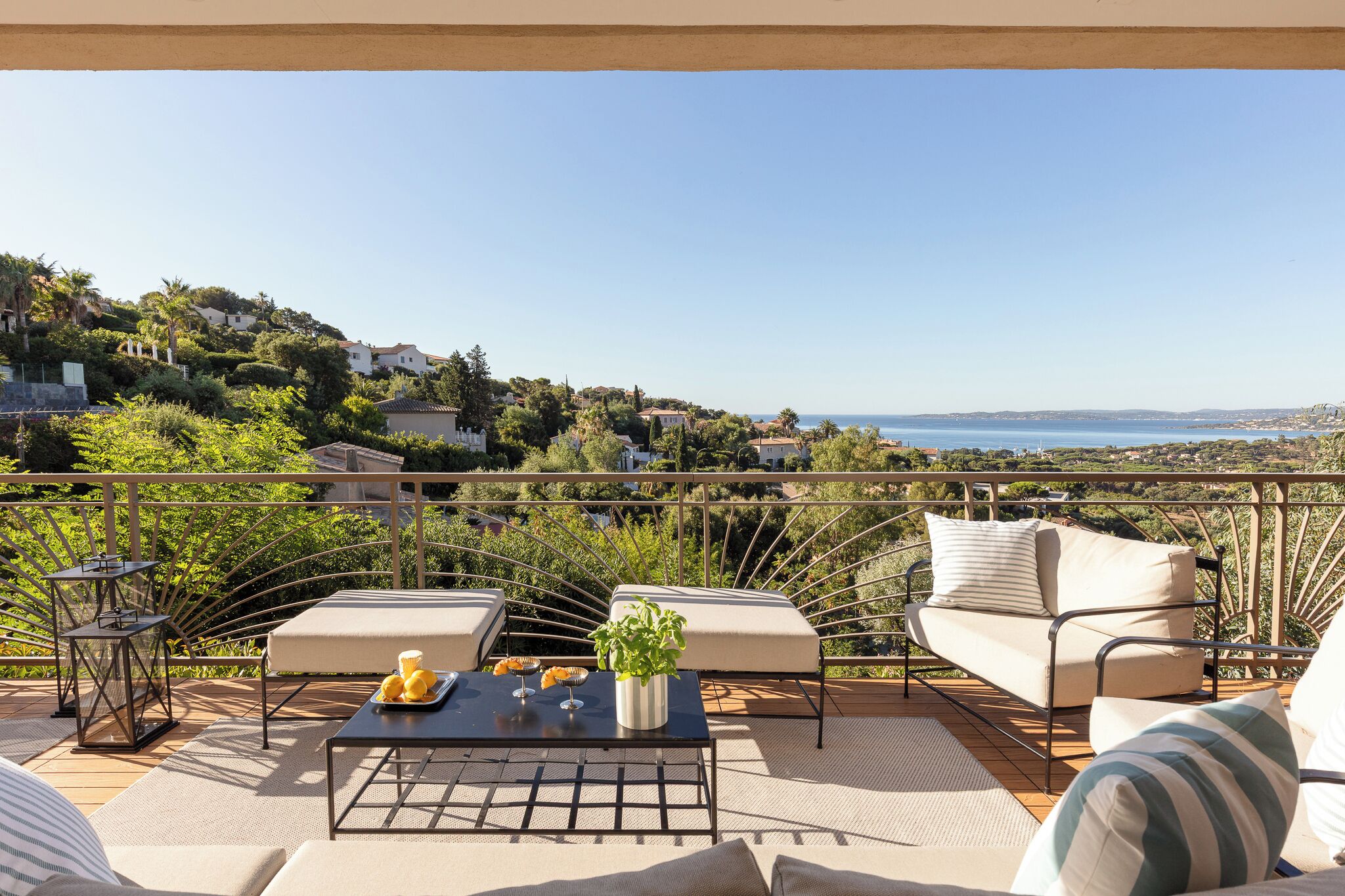 A terrace with outdoor furniture, a table with a plant, and a scenic view of the sea and hills.