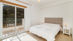 A bedroom with a large bed, a nightstand, a bedside table, and a sliding glass door leading to a balcony.
