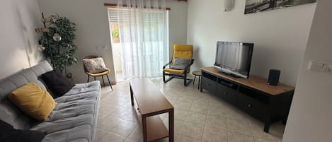A living room with a grey sofa, yellow and black cushions, a wooden coffee table, a TV on a wooden stand, a small round table, and a chair.