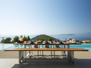 Et spisebord dækket med forskelligt bestik og glas, med udsigt til en pool og et smukt landskab med hav og bakker.