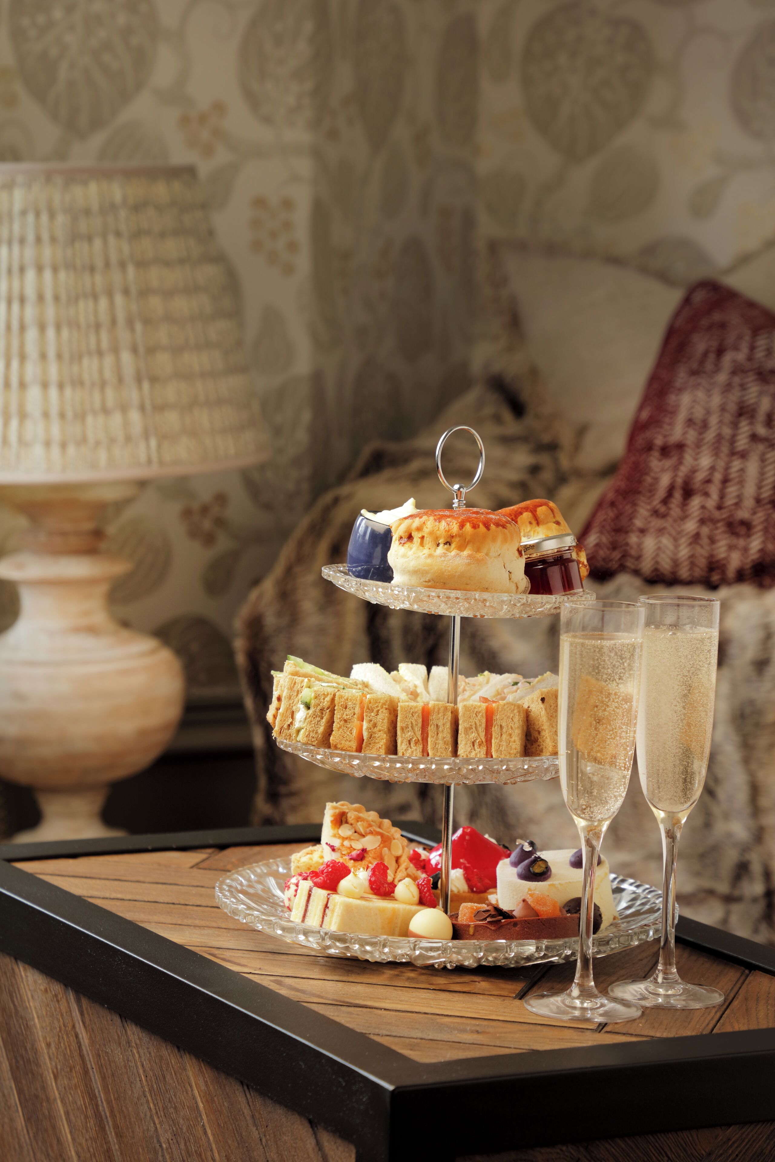 Een drielaags etagère met diverse gebakjes en sandwiches, vergezeld van twee glazen champagne, op een houten tafel met een lamp op de achtergrond.