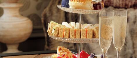 Een drielaags etagère met diverse gebakjes en sandwiches, vergezeld van twee glazen champagne, op een houten tafel met een lamp op de achtergrond.