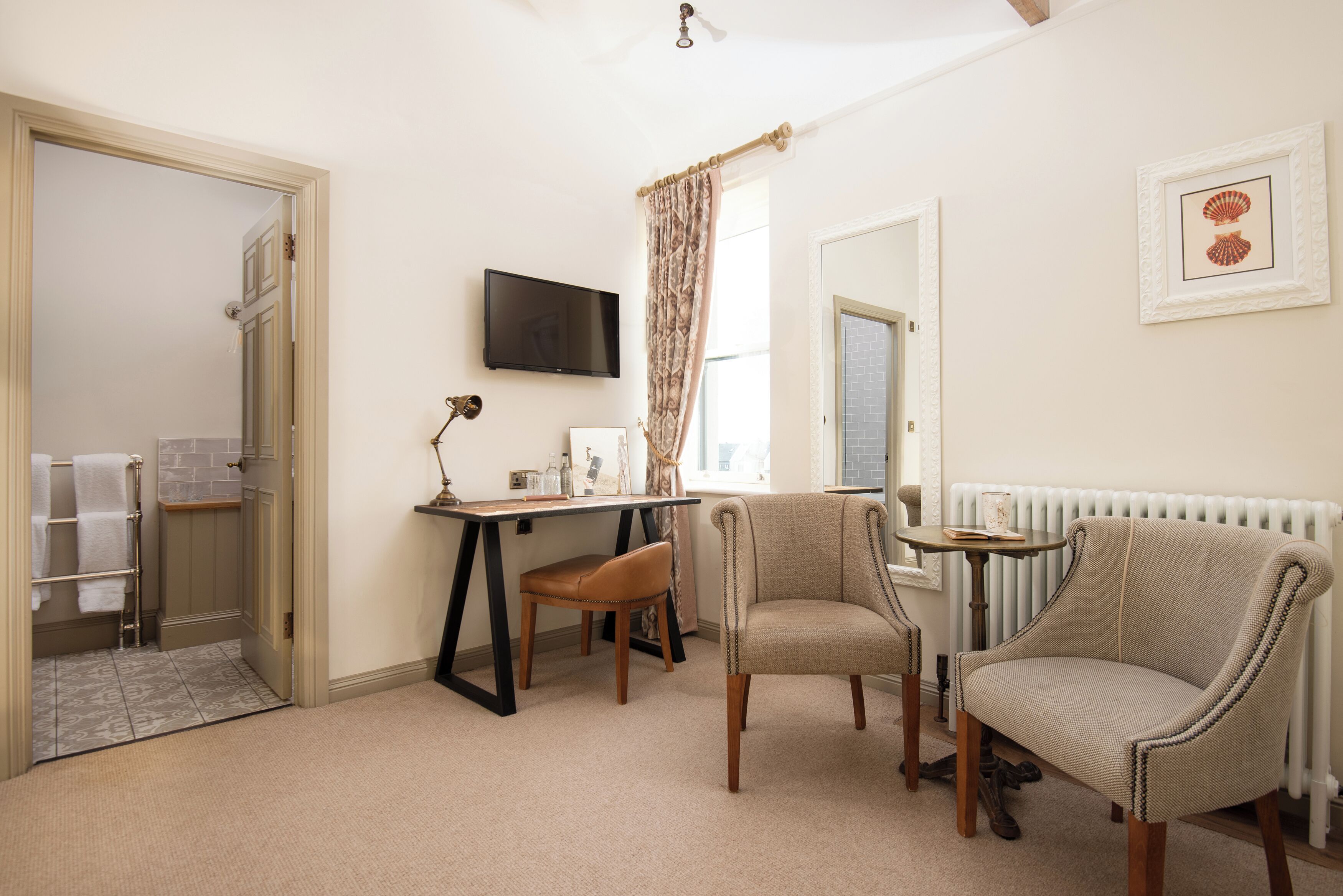 A room with a desk, two chairs, a television, a radiator, and a door leading to a bathroom.