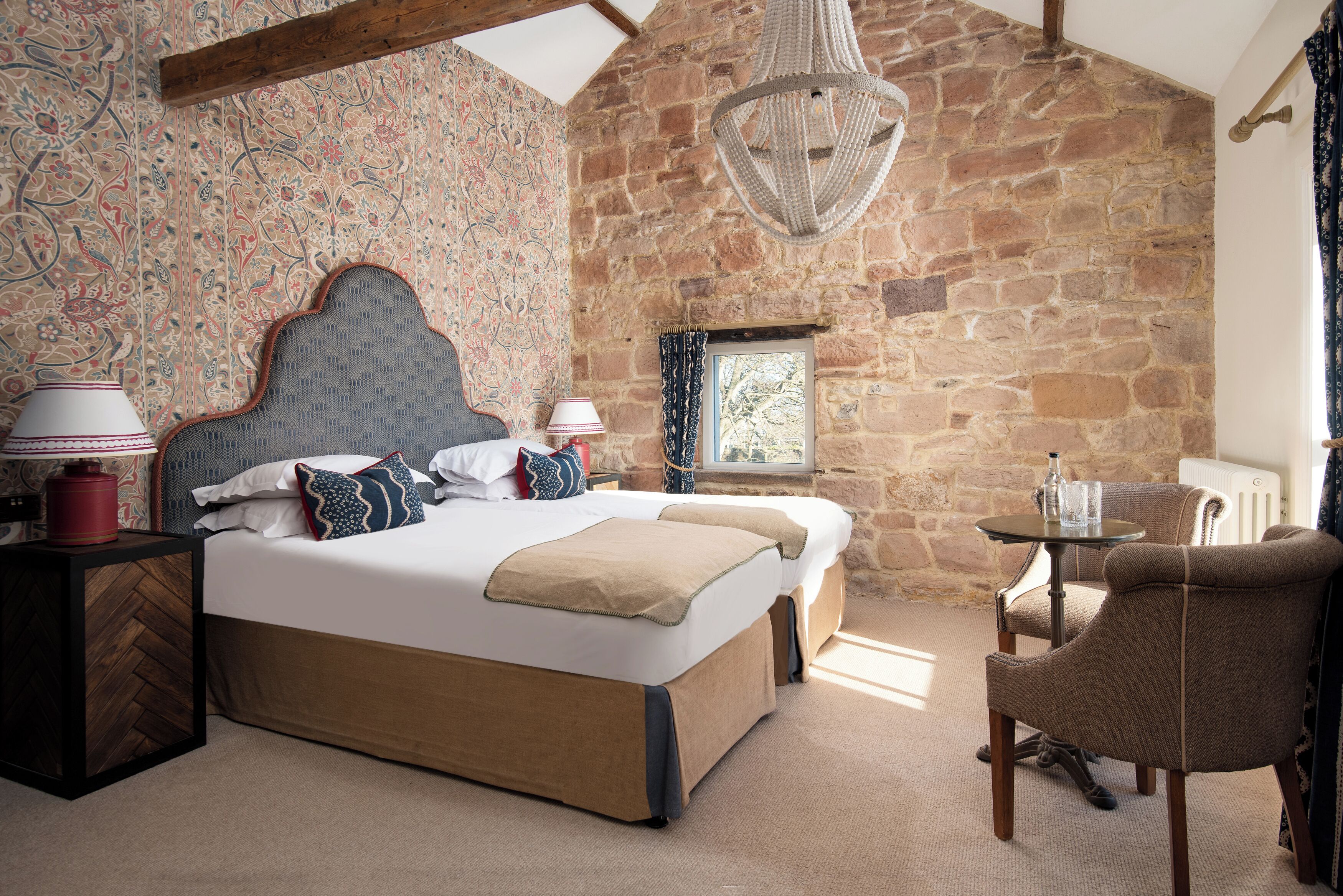 A bedroom with a large bed, a side table, a chair, a window, and a stone wall.