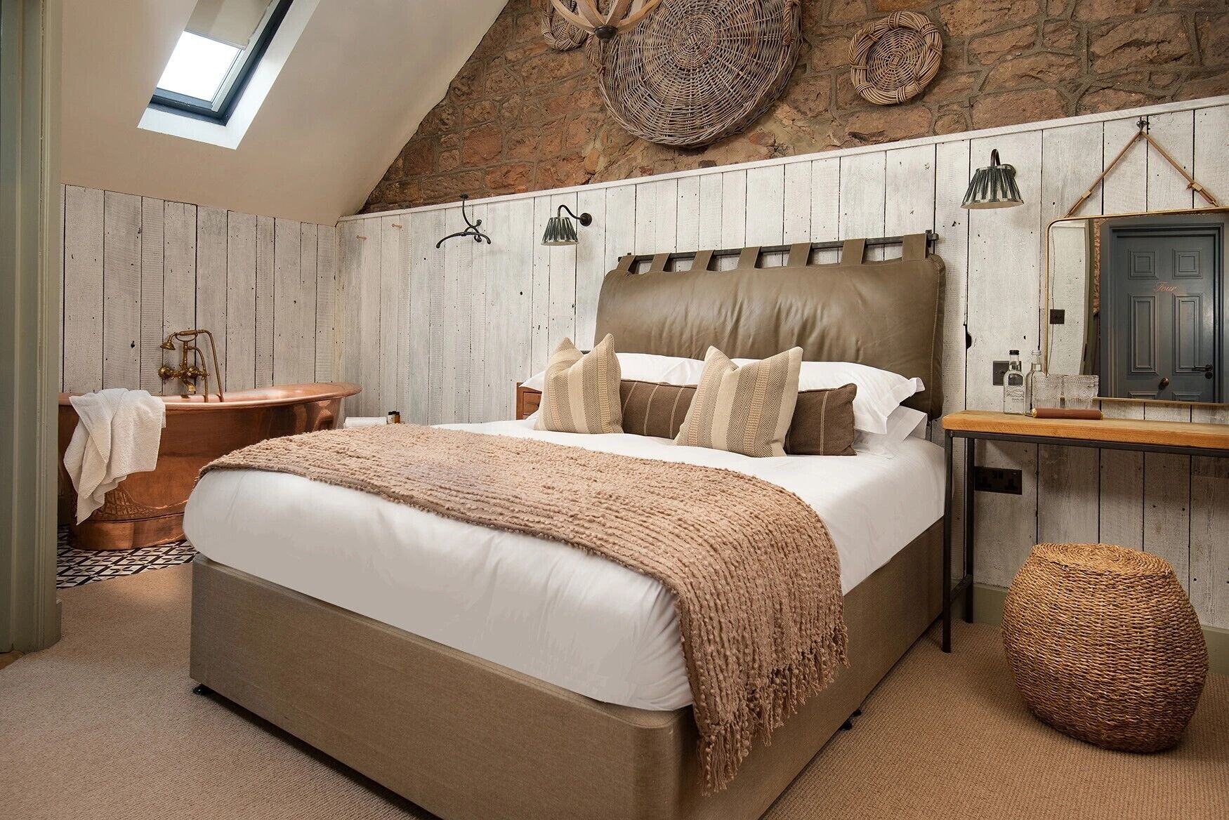 A bedroom with a large bed, a wooden headboard, a small table, a woven stool, and a stone wall with a skylight.