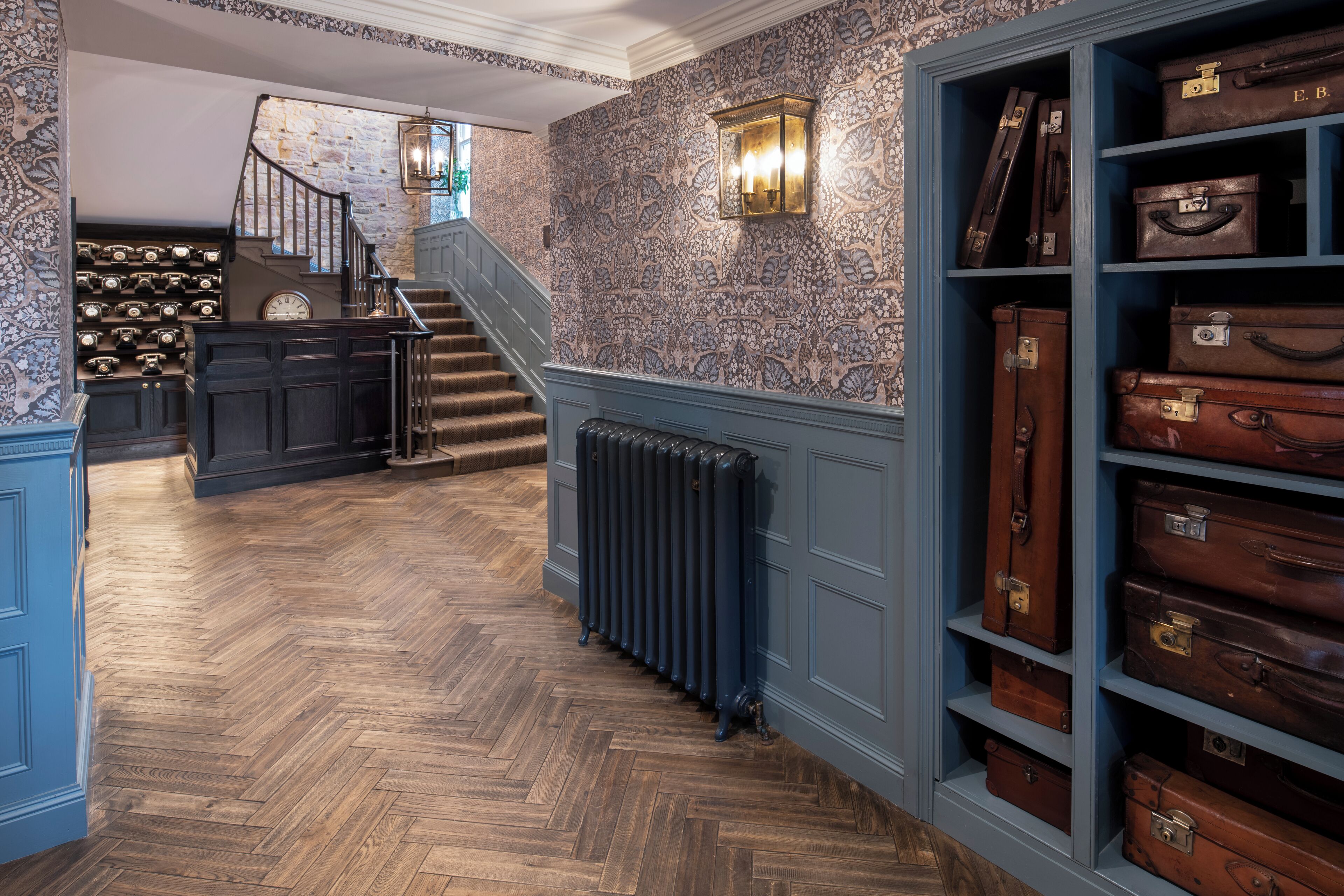 A hallway with patterned wallpaper, a wooden herringbone floor, a staircase, and a wall of suitcases.