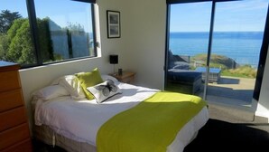 Een slaapkamer met een bed, nachtkastje, dressoir en een groot raam met uitzicht op zee en kliffen.
