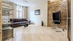 Un salon moderne avec un plancher en bois, un canapé gris, une table basse en verre, un téléviseur fixé sur un mur en briques et une étagère en métal sur laquelle se trouve un chat.