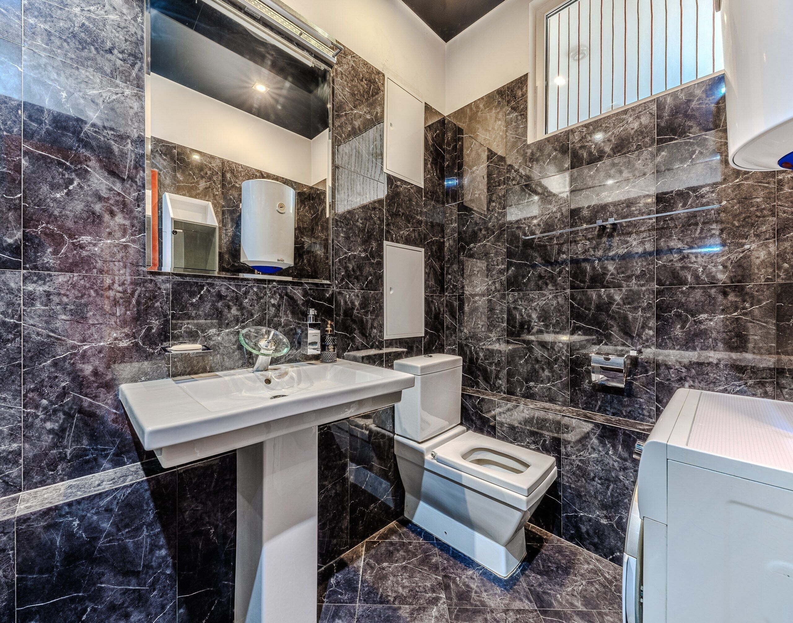 A modern bathroom with a white sink, toilet, and shower area, featuring dark marble tiles and a glass shower enclosure.