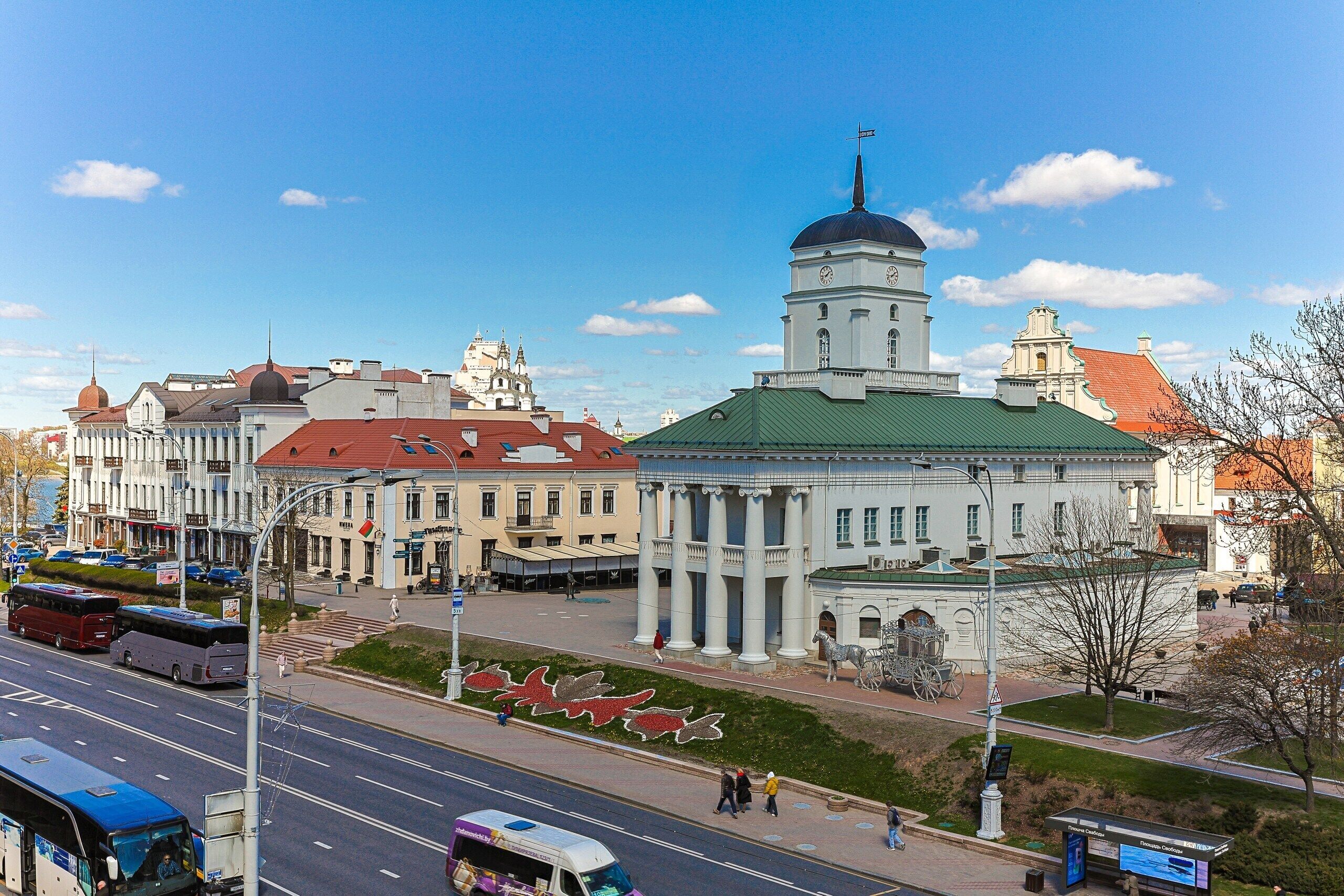 A historic building with a prominent tower and columns, situated next to a wide street with buses.