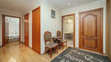 A hotel hallway with wooden doors, a dining area with a table and chairs, and a bed visible in the background.