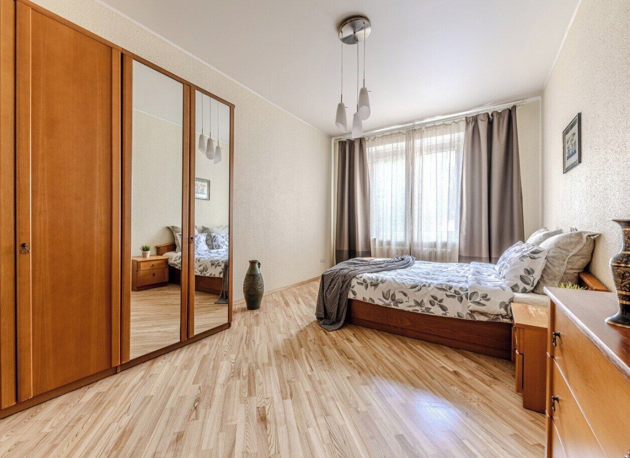 A bedroom with a wooden wardrobe, a bed with patterned bedding, a bedside table, a vase, and a window with curtains.