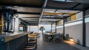 Uma área de refeições ao ar livre com uma mesa comprida e cadeiras, um balcão de bar e vista para o oceano.