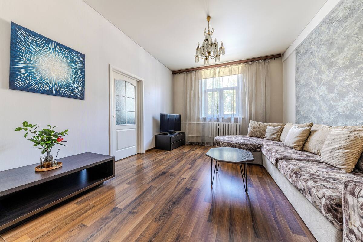 A modern living room with a wooden floor, a sectional sofa, a flat-screen TV, a decorative plant, and a large abstract painting.