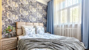 A bedroom with a cityscape wallpaper, a bed with a floral duvet, a bedside table with a vase, and a window with sheer curtains. - StudioMinsk Apartments in Centre (Minsk)