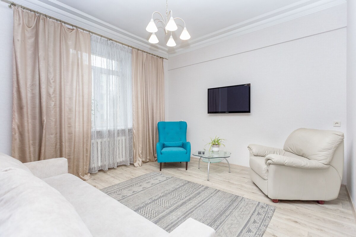 A modern living room with a beige sofa, a blue armchair, a beige armchair, a small glass table with a plant, a TV mounted on the wall, and a patterned rug.
