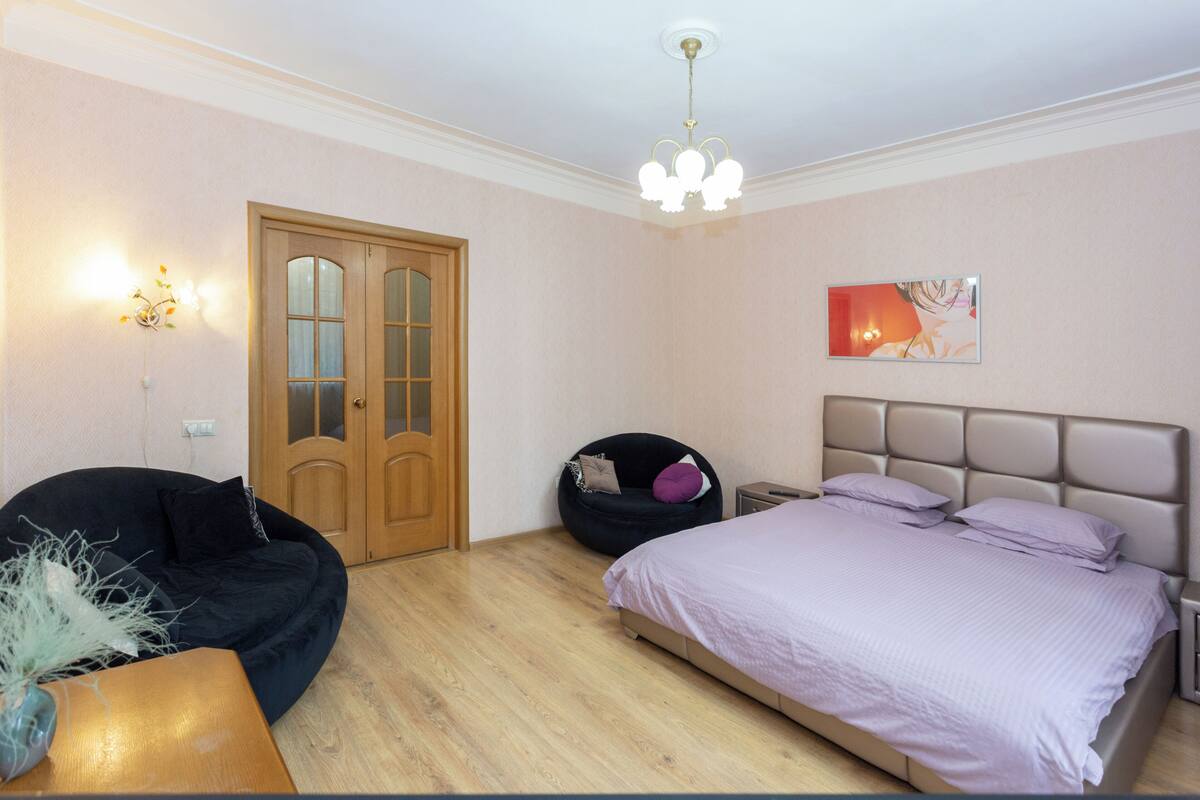 A bedroom with a bed, a chair, a door, and a wall-mounted light fixture.