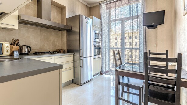 A modern kitchen with stainless steel appliances, a dining area with a wooden table and chairs, and a large window with a view of buildings. - StudioMinsk Apartments in Centre (Minsk)