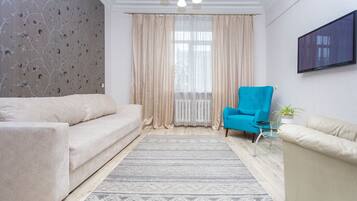 A living room with a beige sofa, a blue armchair, a glass coffee table, and a TV on the wall.