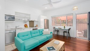 A modern kitchen with white cabinets, a microwave, and a refrigerator. There is a teal sofa and a wooden coffee table with a book. A dining area with a table and chairs is visible through a window. - Yamba Daze Sunset - Walk to the pet-friendly beach from your pet-friendly stay (Yamba)