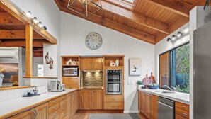 Eine moderne Küche mit Holzschränken, Edelstahlgeräten, einer großen Uhr und Blick ins Freie.