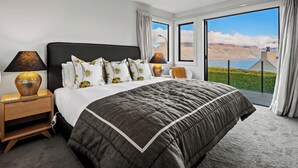 Ein modernes Schlafzimmer mit einem großen Bett, zwei Nachttischen, einer Lampe und Blick auf eine gebirgige Landschaft durch große Fenster.