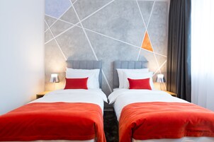 A hotel room with two beds, each with a red blanket, white pillows, and a grey headboard. There are bedside lamps and a geometric wall design.
