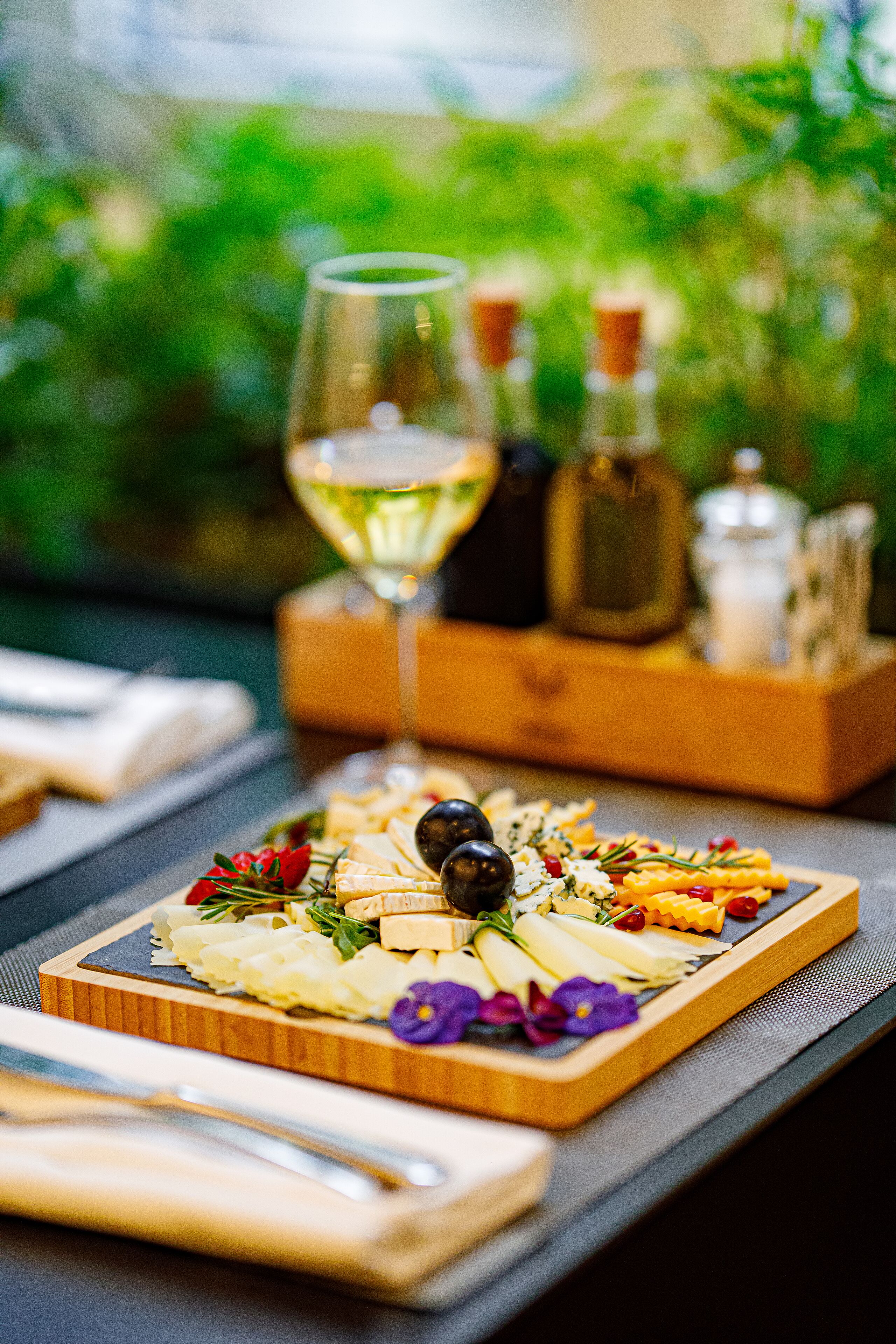 A wooden board with a variety of cheeses, olives, and flowers, accompanied by a glass of white wine and bottles of olive oil.