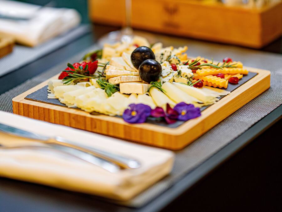 A wooden board with a variety of cheeses, olives, and flowers, accompanied by a glass of white wine and bottles of olive oil.