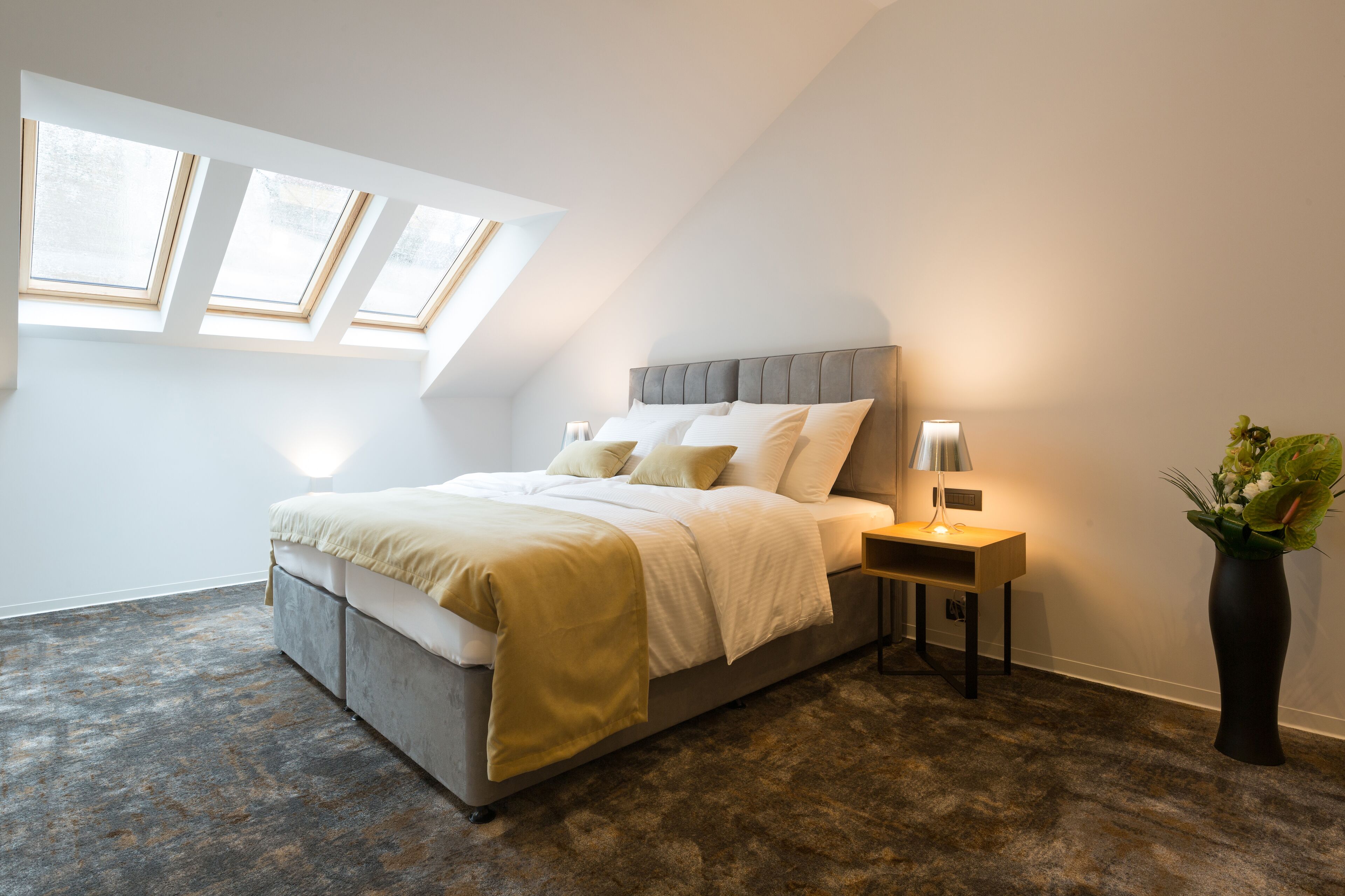 A bedroom with a bed, bedside table, lamp, and a vase with flowers.
