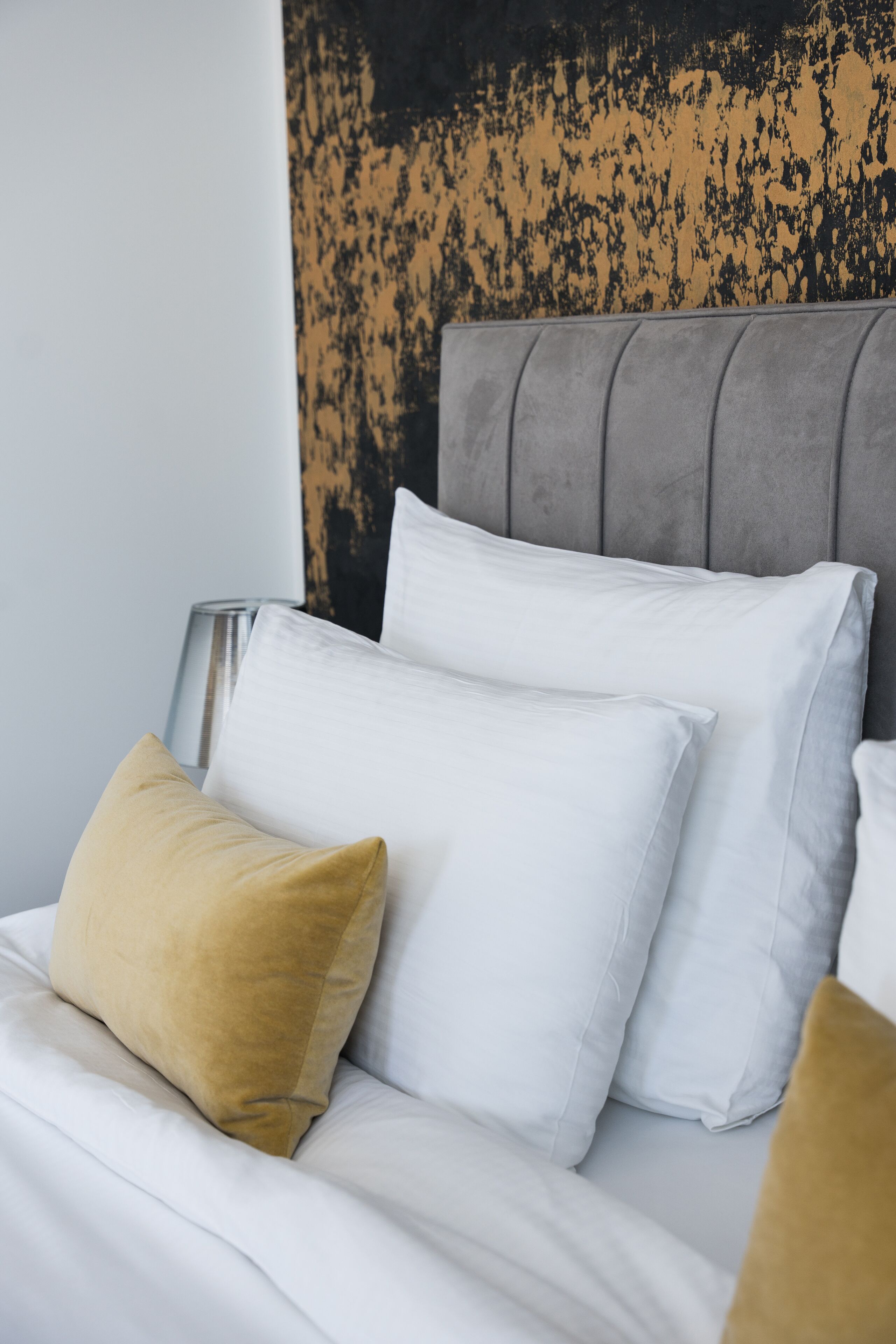A bed with white and mustard pillows, a gray headboard, and a textured wall behind.