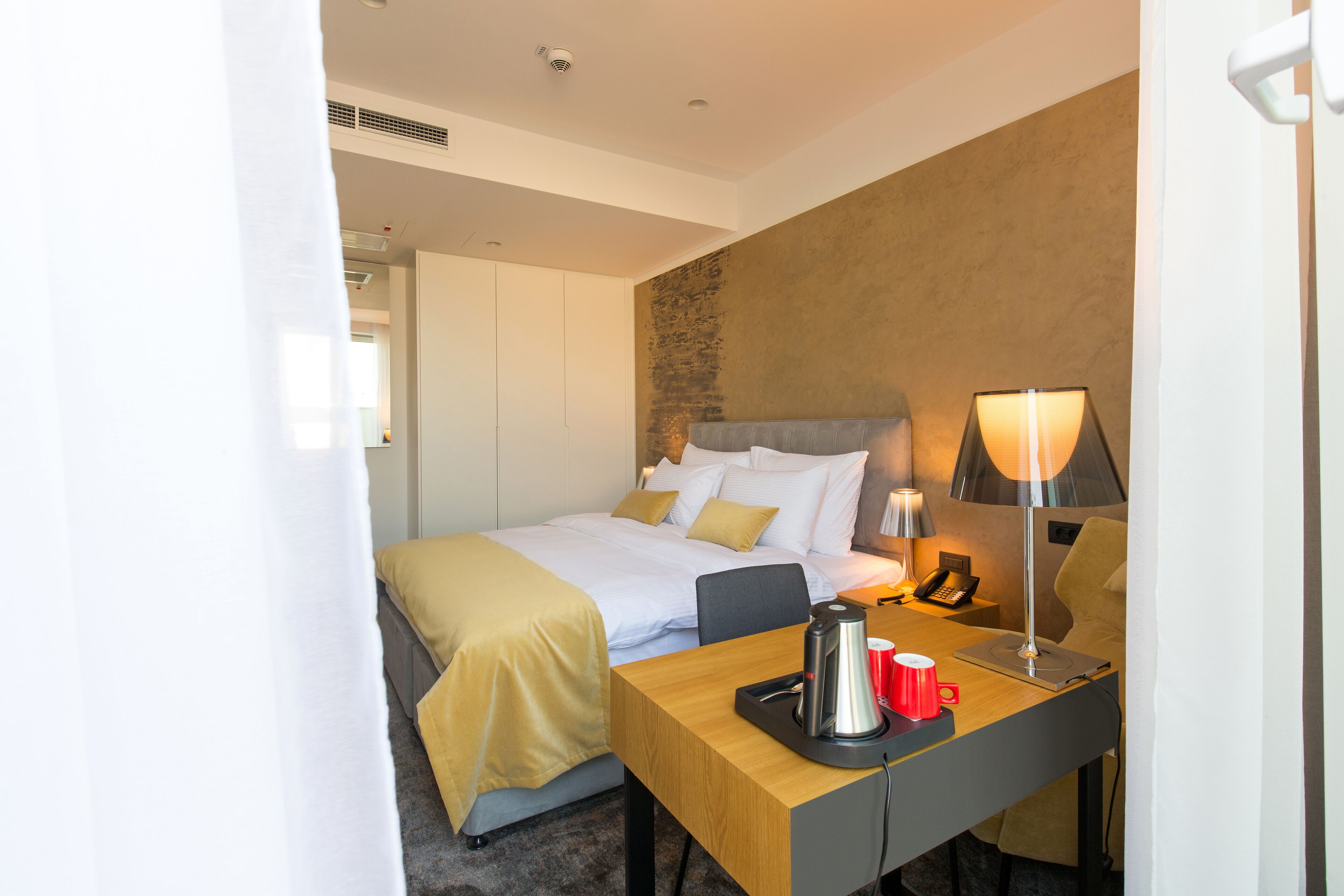A hotel room with a bed, bedside table with a lamp, a telephone, and a red mug.