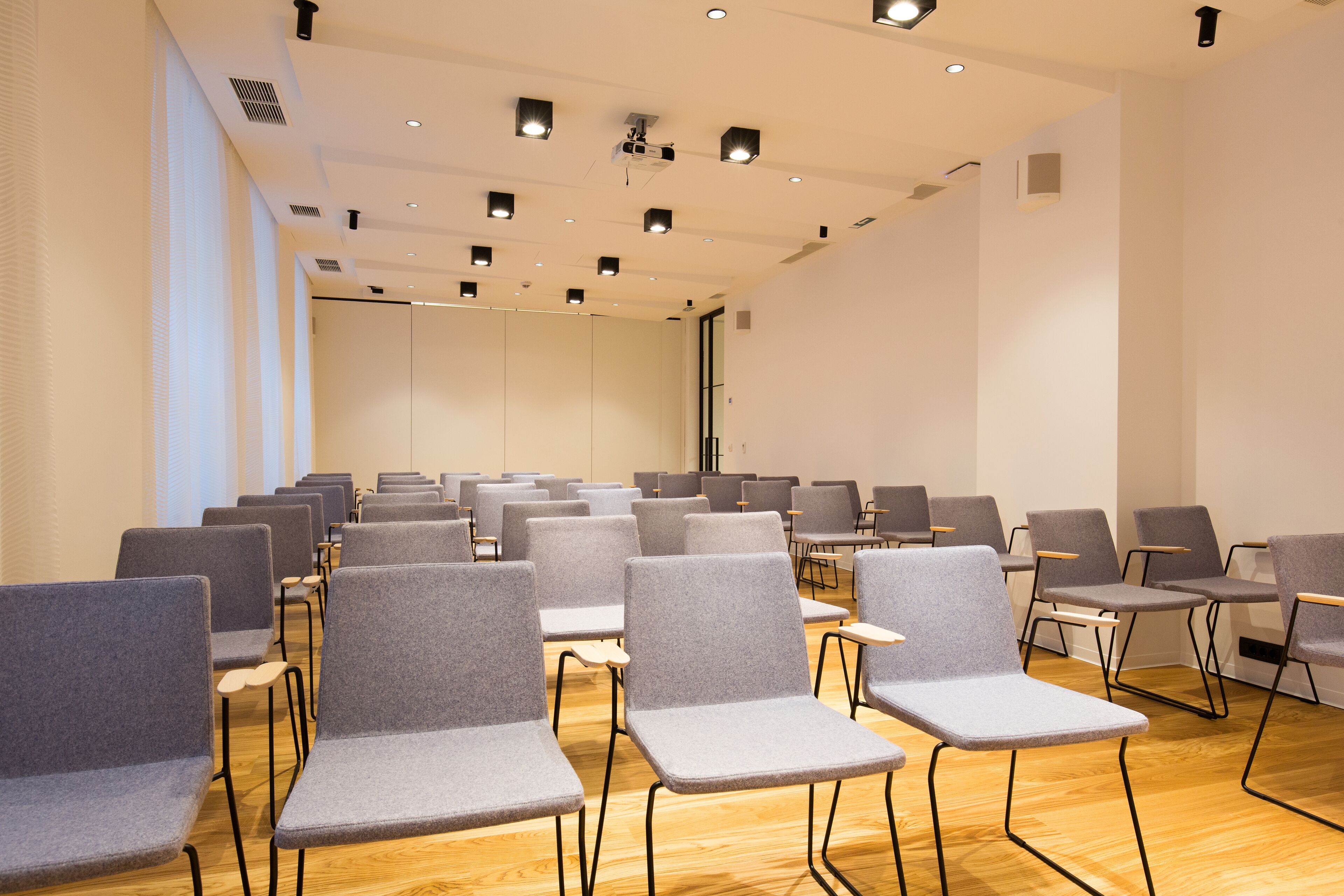 A conference room with rows of grey chairs, wooden floors, and recessed ceiling lighting.