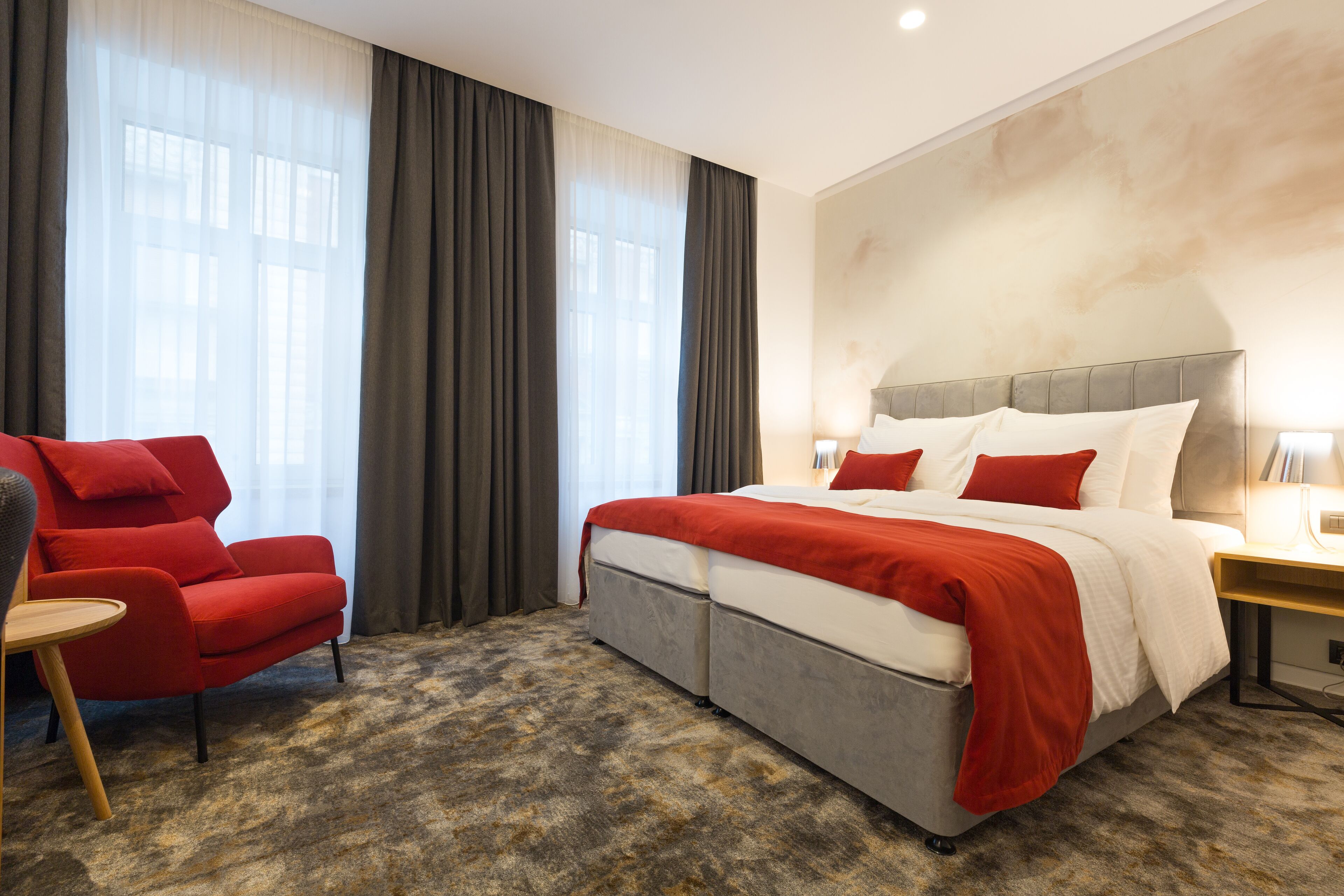 Quarto de hotel com uma cama grande, uma poltrona vermelha e piso carpeteado.