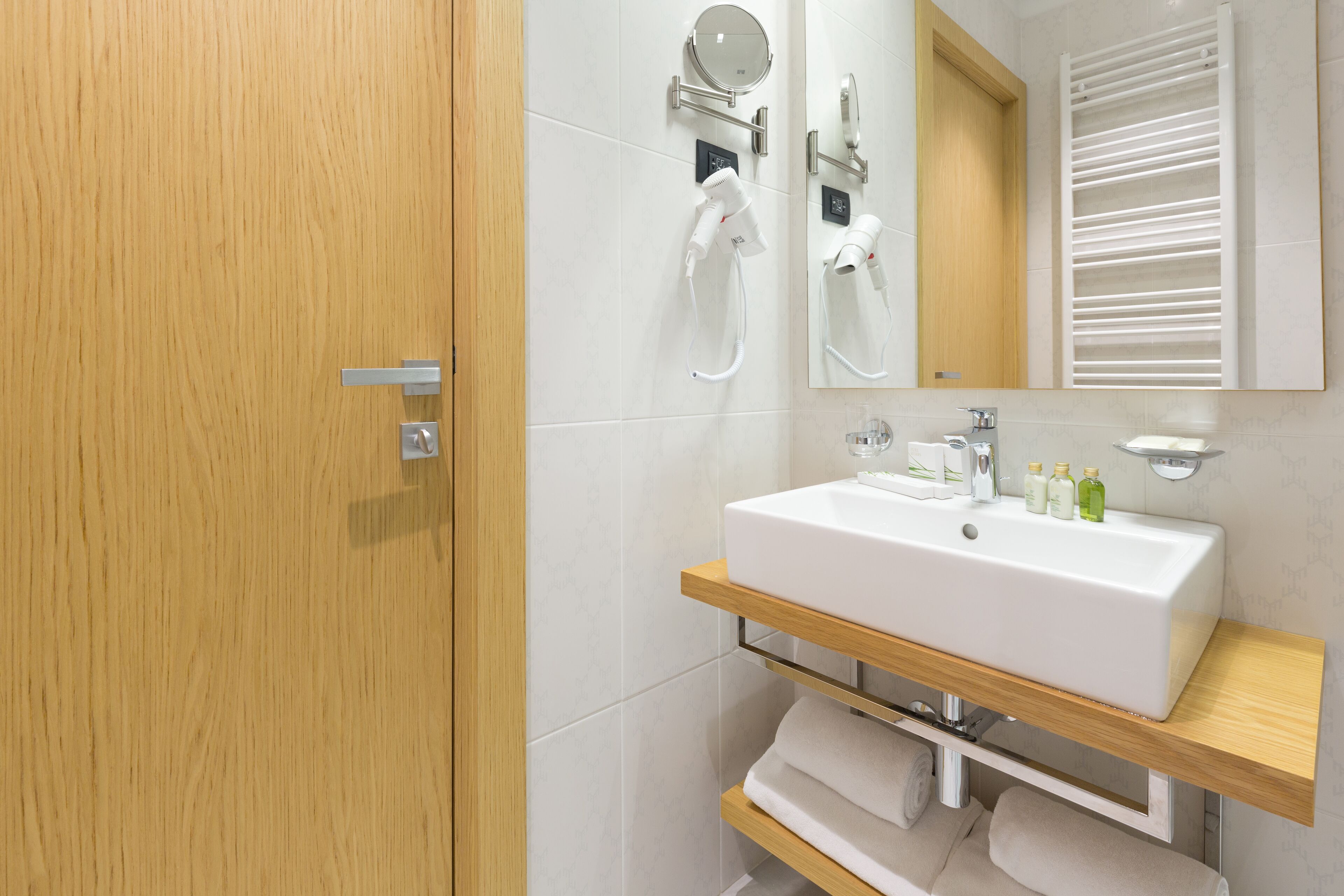 A modern bathroom with a wooden door, white tiles, a large mirror, a white sink, and a towel rack.