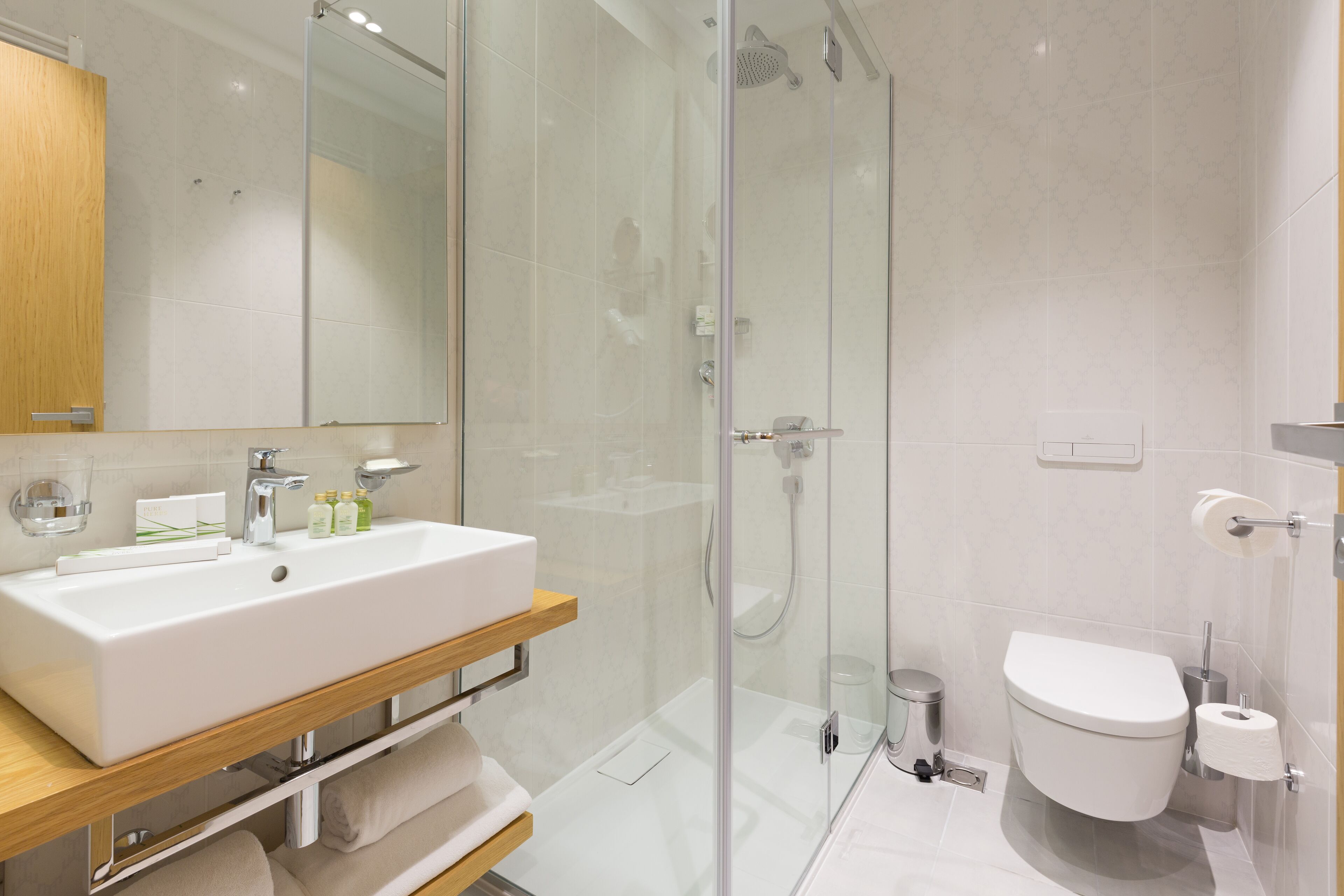 A modern bathroom with a glass-enclosed shower, a white sink with a wooden vanity, and a toilet with a paper towel holder.