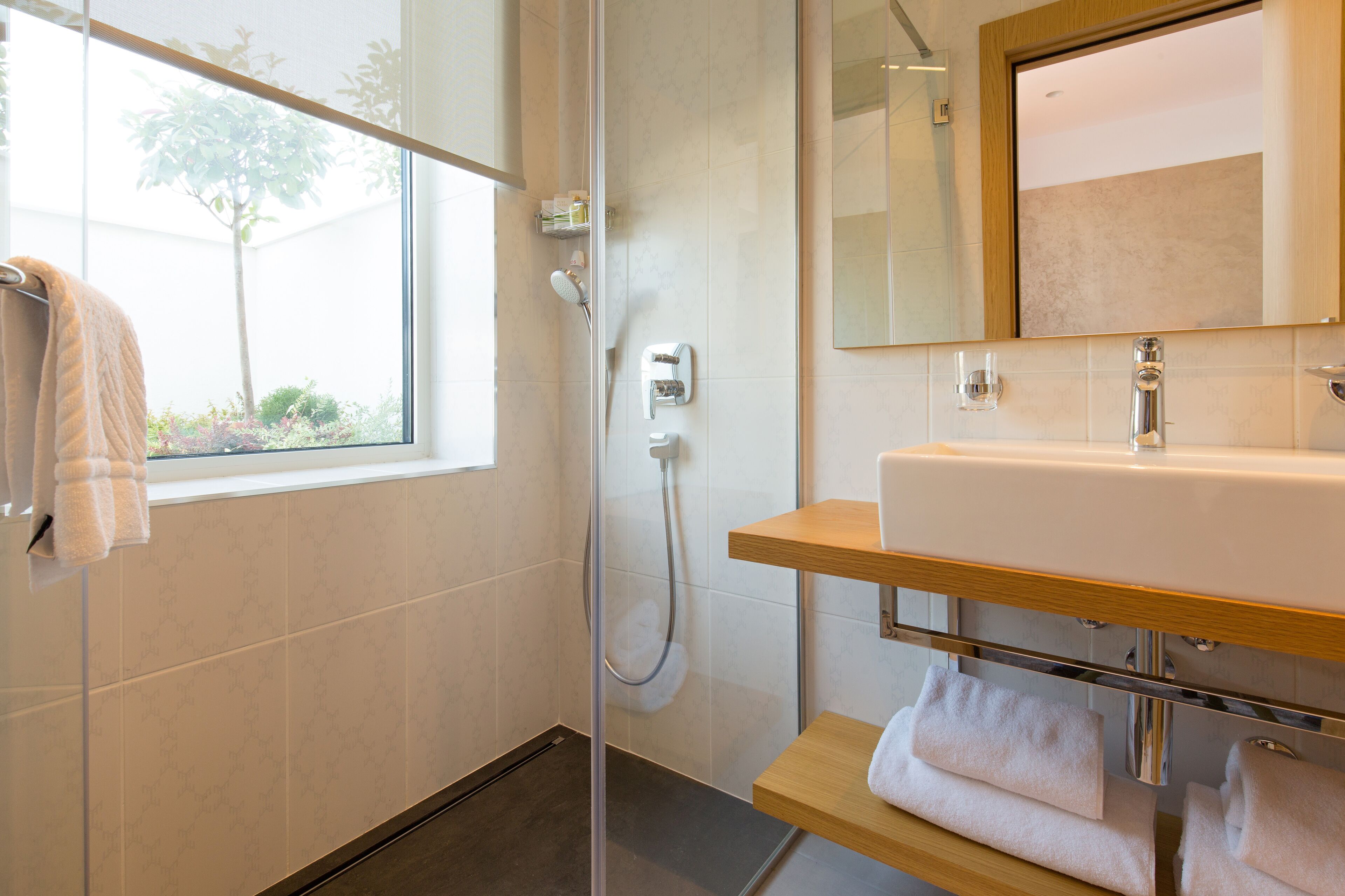 Une salle de bain moderne avec une douche à l’italienne en verre, un lavabo blanc doté d’une étagère en bois et une fenêtre sur laquelle est accrochée une serviette.
