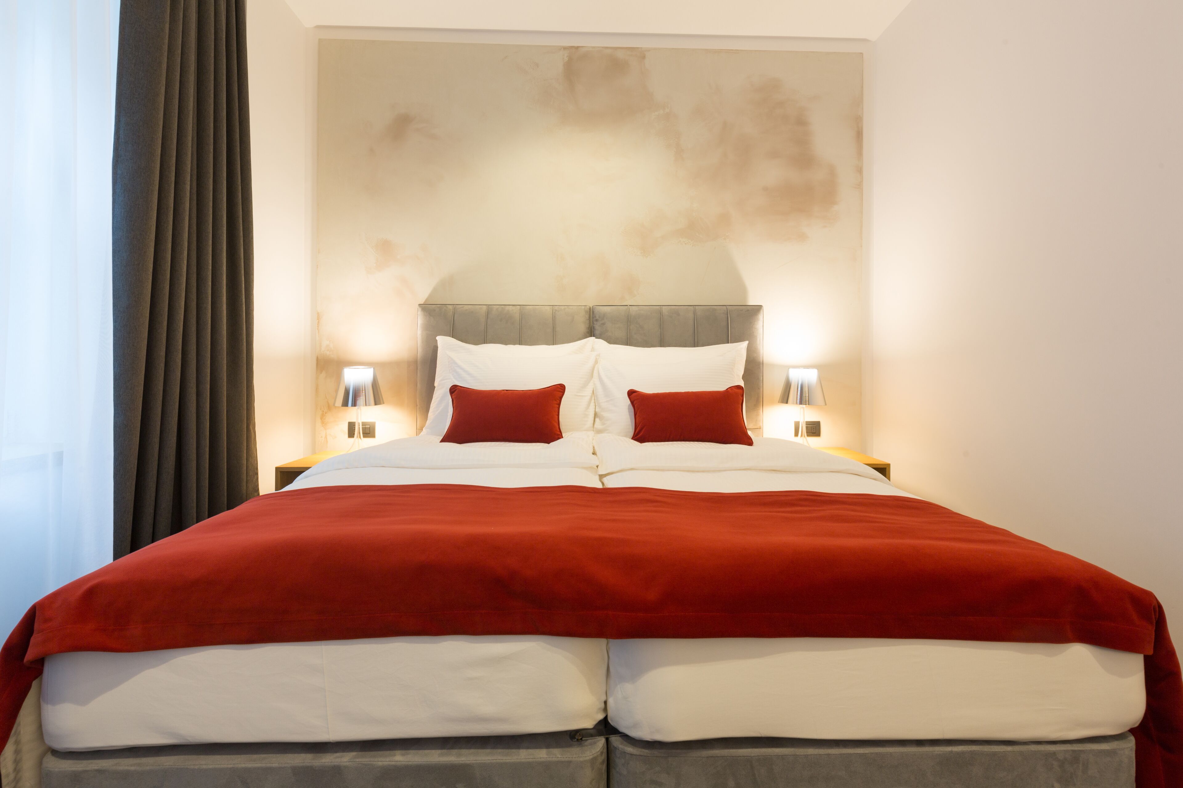 A double bed with a red and white bedspread, two red pillows, and a headboard with a textured wall behind it.