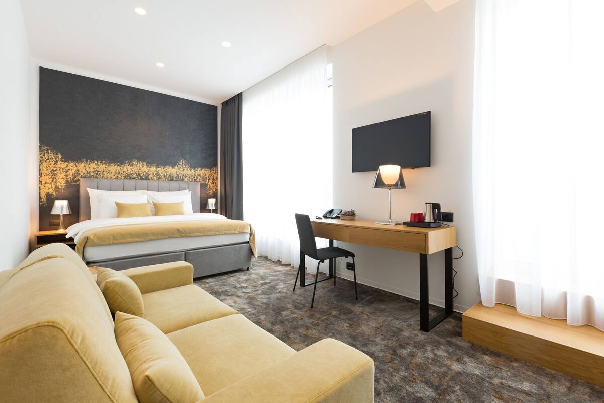 A modern hotel room with a bed, a desk, a chair, a sofa, and a large artwork on the wall.