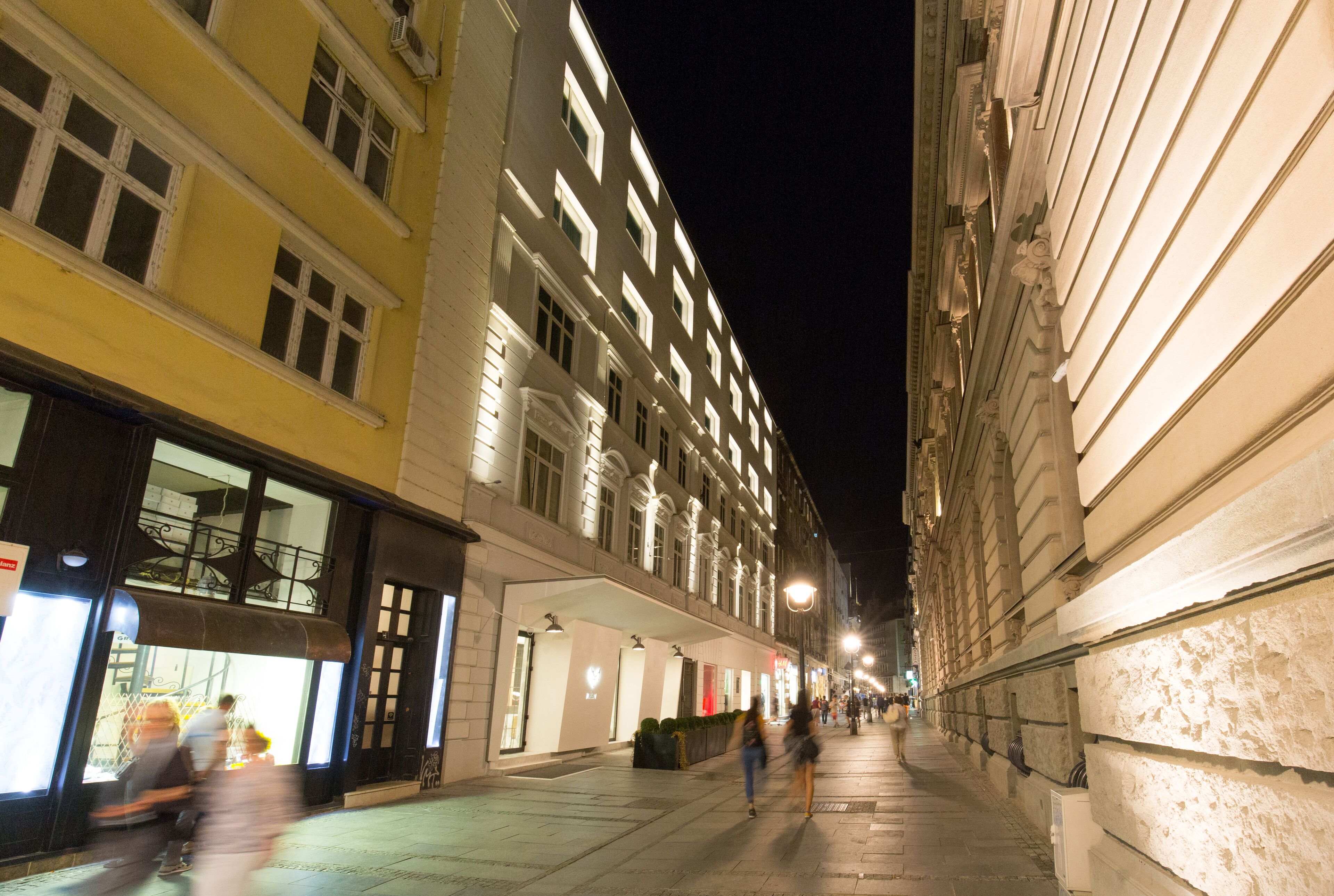 Vista nocturna de una calle bordeada por edificios de varios pisos, con escaparates iluminados y una acera para peatones.
