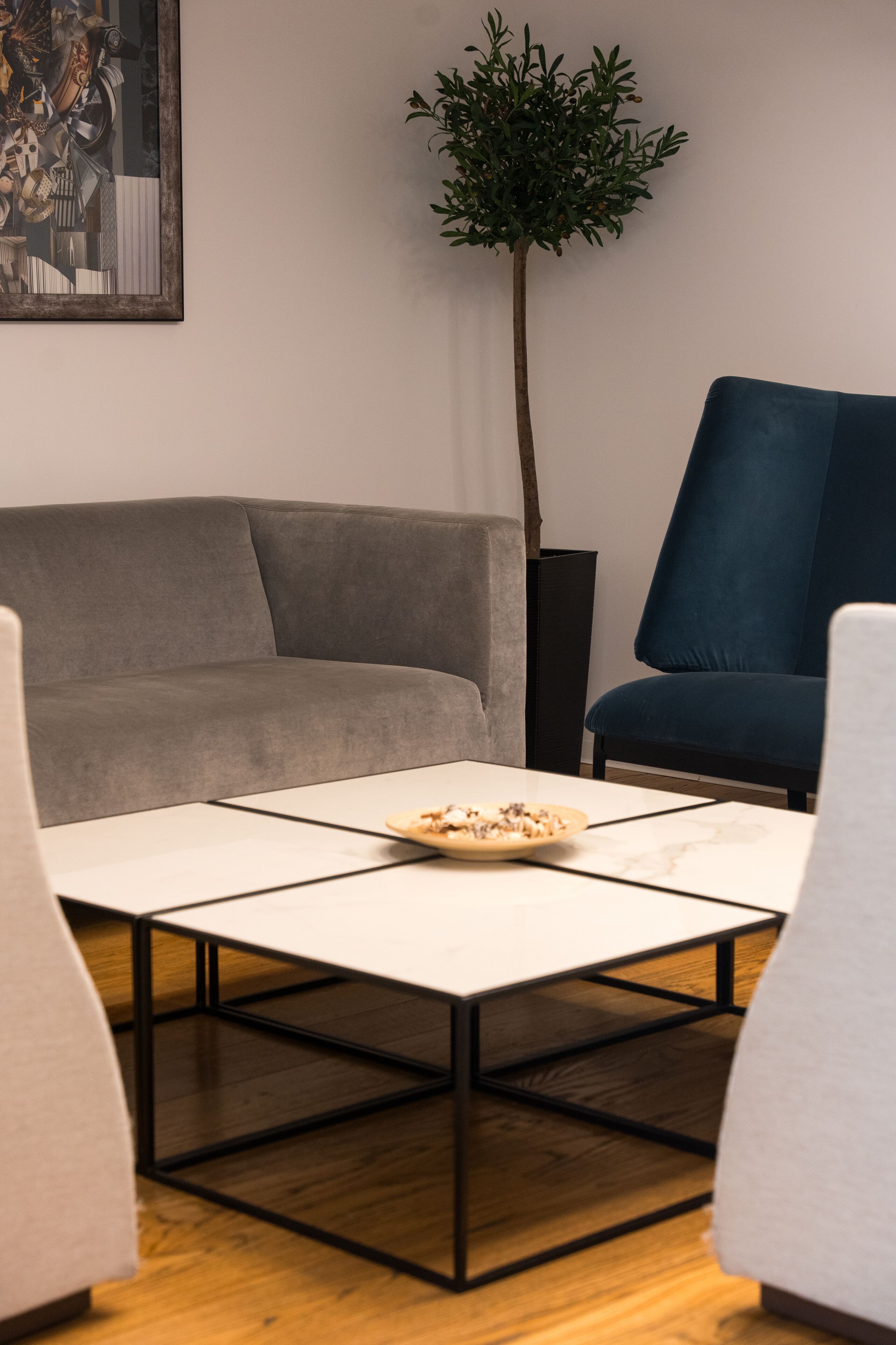 A modern living room with a grey sofa, a blue chair, a white coffee table with a bowl of cereal, and a tall green plant.