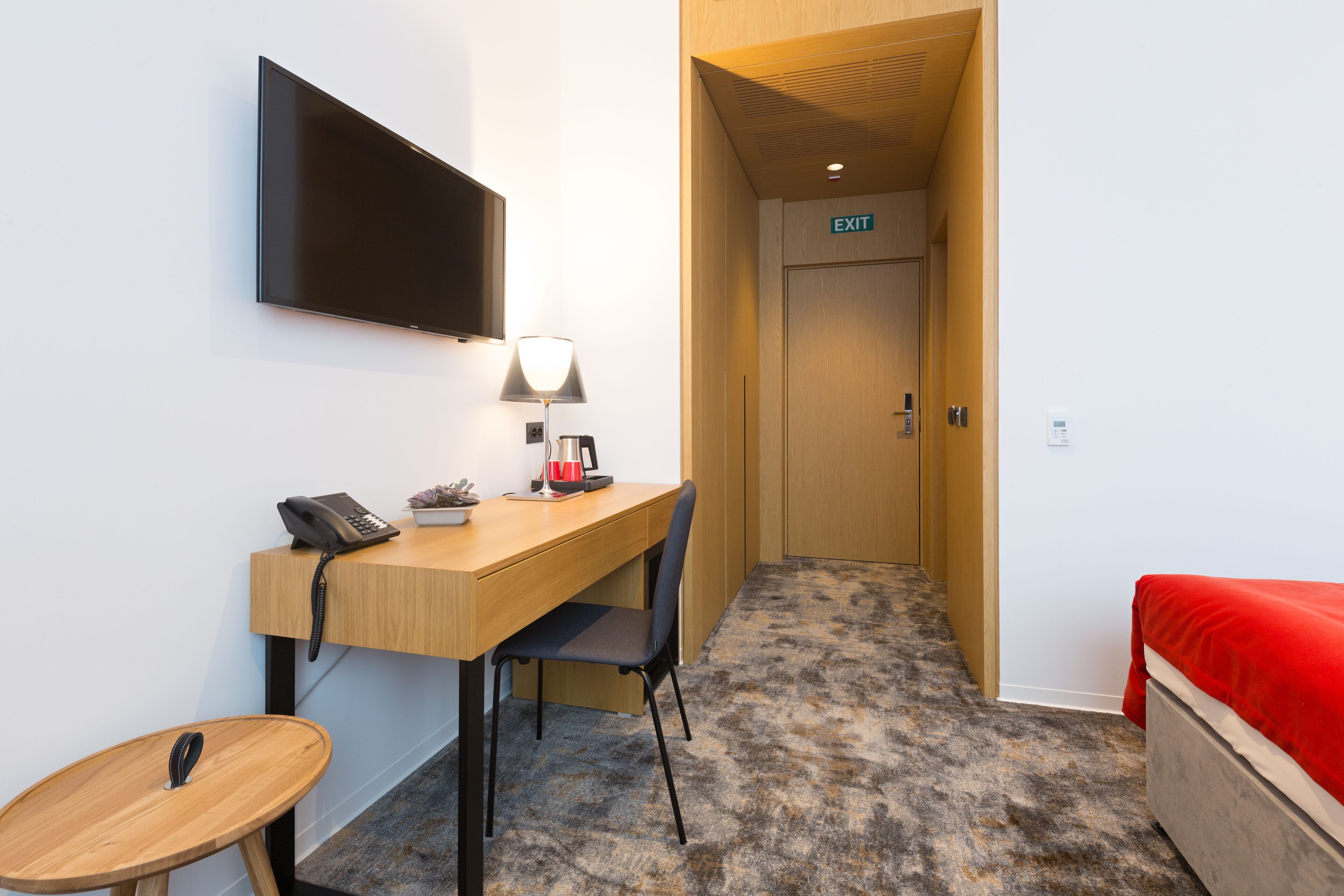 Une chambre d’hôtel avec un bureau en bois, une chaise, une petite table ronde, un lit recouvert d’une couverture rouge, un téléviseur fixé au mur et un panneau indiquant la sortie.