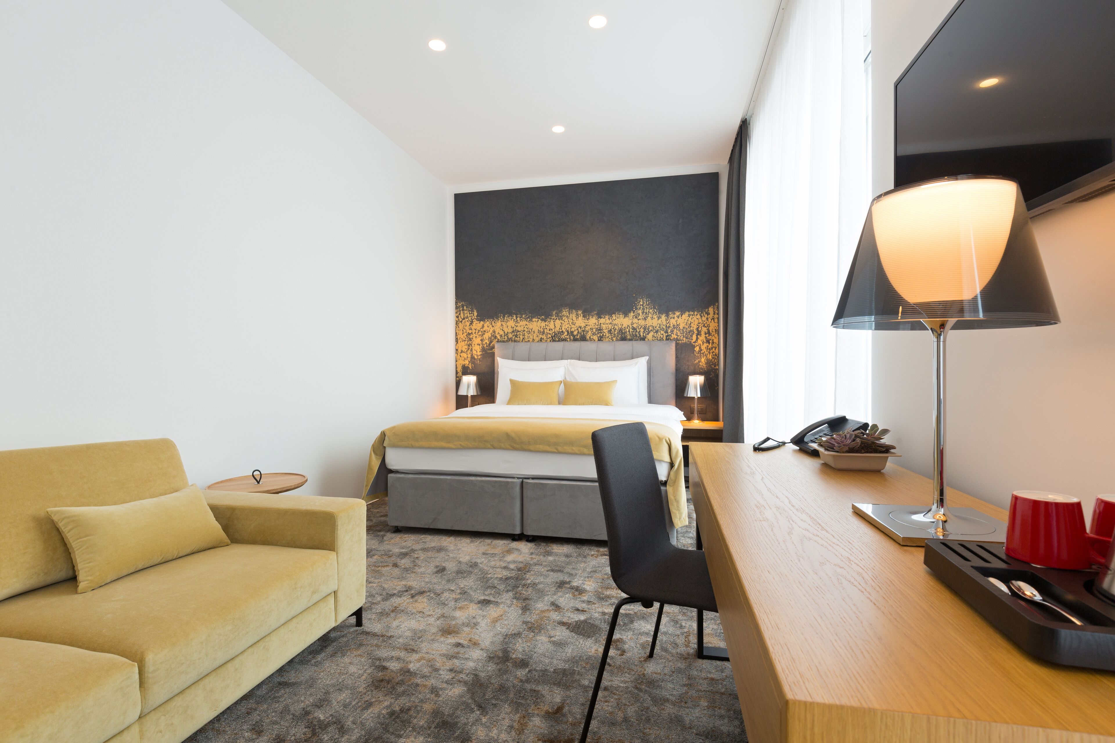 A modern hotel room with a bed, a sofa, a desk, and a lamp.