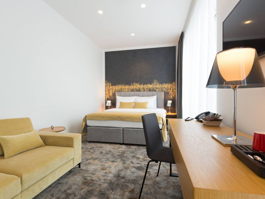A modern hotel room with a bed, a sofa, a desk, and a lamp.